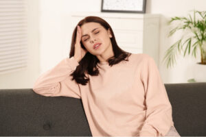 Woman sitting on a sofa with her eyes closed and holding her temple as if experiencing a headache.