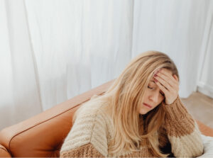 Woman sitting on a couch with her hand on her forehead while experiencing fatigue and widespread discomfort.