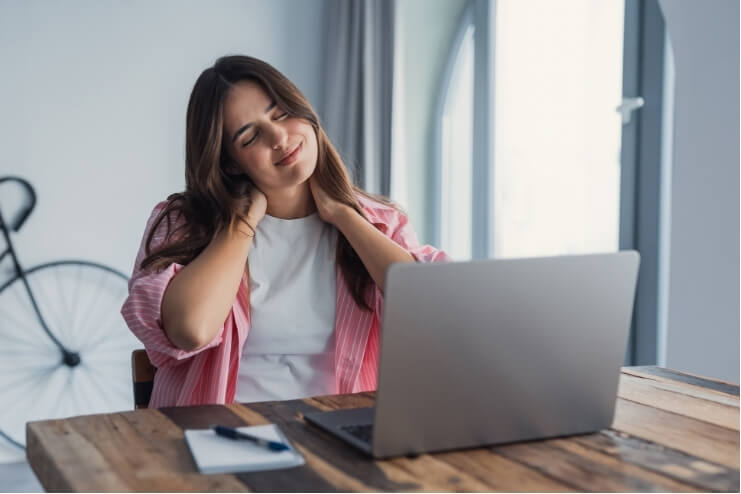 Woman sitting at a laptop holding her neck, representing neck headaches related to desk work and poor neck control.