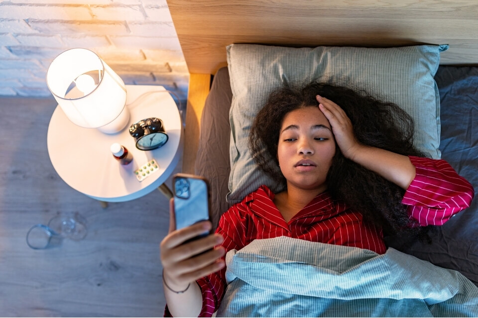 Woman lying awake in bed with one hand on her forehead, looking at her phone while experiencing migraine symptoms.