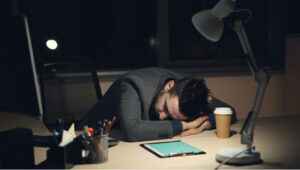 Man asleep with his head resting on his arms at a desk beside a coffee cup and lamp.