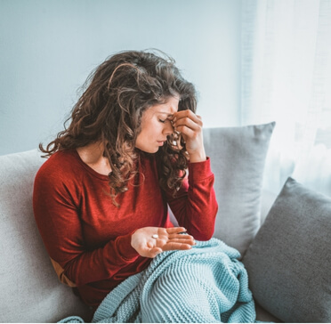 Woman sitting on a couch with headache, holding medication in one hand and touching her forehead