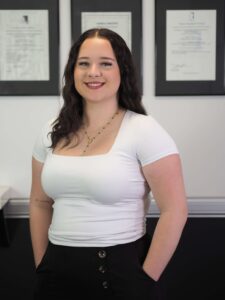 Holly Davies, Administrator at Brisbane Headache and Migraine Clinic, smiling in a white top with black trousers, standing in front of framed certificates.